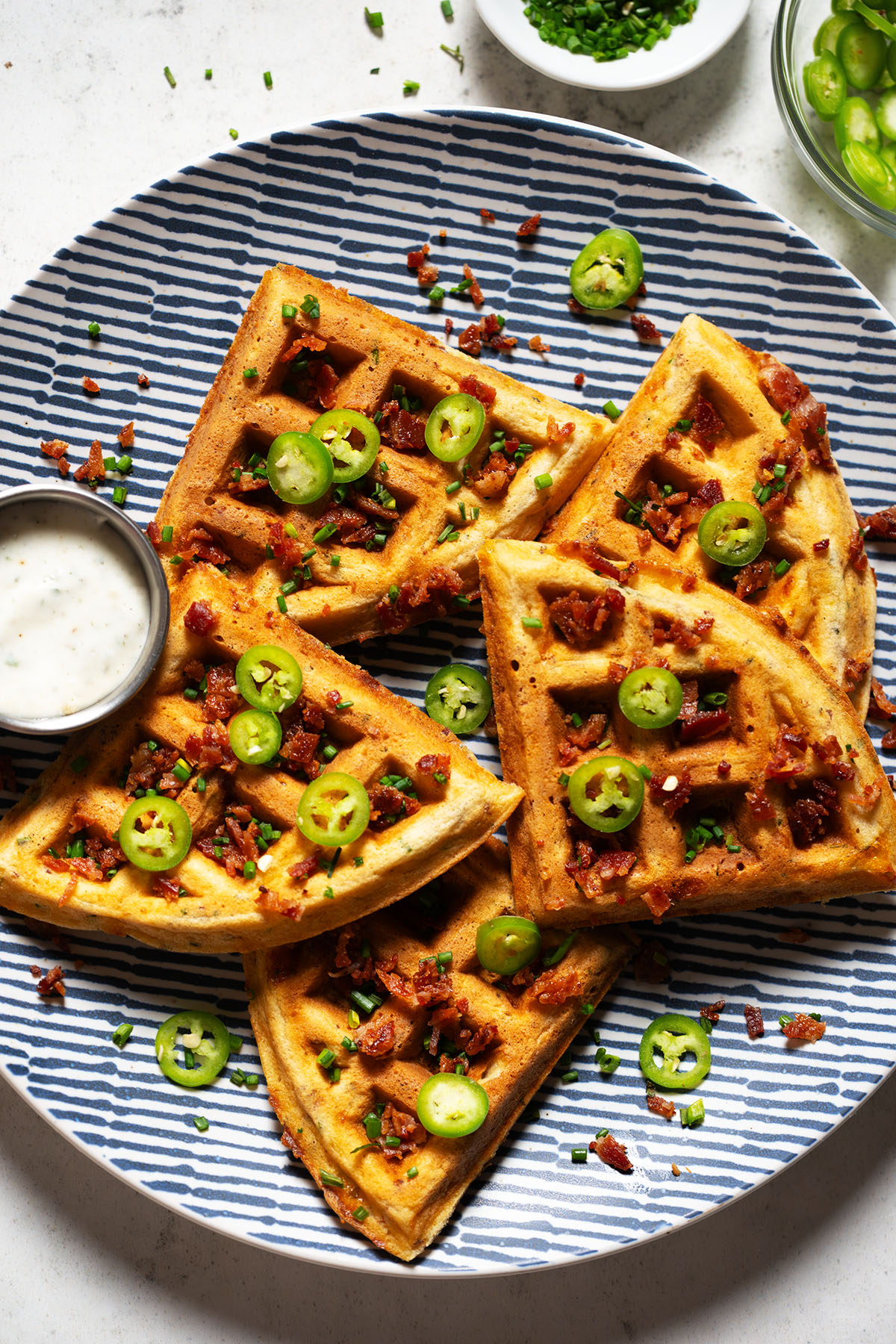 Crispy bacon cheddar savory waffles topped with jalapeño slices and chives, served with ranch dressing on a blue striped plate.